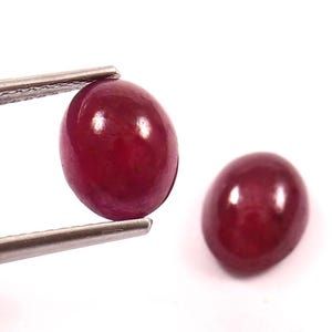 May include: Two polished, oval-shaped, deep red gemstones. One is held by a metal tweezer, the other rests freely. The gems have a smooth, reflective surface, and are set against a bright white background.