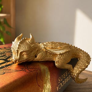 Könnte beinhalten: Eine goldfarbene Drachenfigur mit strukturierten Schuppen, kleinen Flügeln und Hörnern, die auf dem Rand eines Buches ruht. Der Drache ist in einer schlafenden Position dargestellt, mit nach vorne geneigtem Kopf und einem langen, geschwungenen Schwanz.