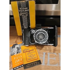 May include: A black Kodak Vigilant Junior Six-20 folding camera with a circular lens assembly. Included is a yellow and black box labeled Kodak Vigilant Junior Six-20, a printed manual, and a Snapshot Kodaguide exposure reference card.