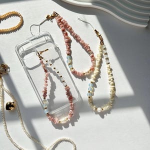 May include: Two phone charms made with pink and white beads. The charms are attached to a clear phone case. The charms are made with small, irregular shaped beads.