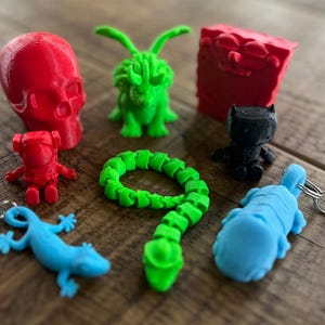 Peut inclure: Une collection de figurines imprimées en 3D, comprenant un crâne rouge, un monstre vert, une figure rouge en forme d'éponge, une figure noire, un astronaute rouge, un serpent vert, un lézard bleu et un chien bleu.