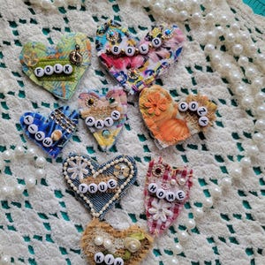 May include: A collection of handmade heart-shaped fabric pins with various patterns and embellishments. Each heart features a different word spelled out with small letter beads, including "FOLK," "BOLD," "TIDE," "PATH," "COZY," "TRUE," "HOME," and "KIN."