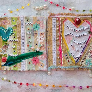 May include: A handmade journal spread with fabric, beads, and text. One page reads "In a field of roses" with a teal heart and green feather. The other page says "Kind heart, brave soul, fierce spirit" in a heart shape.