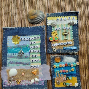 May include: Three denim fabric art pieces with beach-themed embellishments. One says "Sitting on the Dock of the Bay," another "Booze in the Blender," and the third "Somewhere Beyond the Sea." Seashells and charms are included.