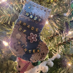 May include: A handmade Christmas stocking ornament with the text "Hazy Shade of Winter." The ornament is made of burlap, denim, and lace, with floral and bead embellishments. It hangs on a Christmas tree.