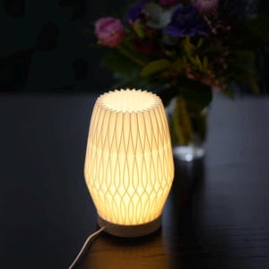 Könnte beinhalten: Eine weiße Tischlampe aus Keramik mit einem geometrisch gemusterten Schirm. Die Lampe ist eingeschaltet und beleuchtet den Raum.