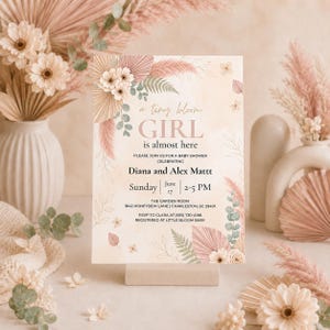 Puede incluir: Una tarjeta de invitación rectangular para baby shower con ilustraciones en acuarela de hojas de palma secas, hierba de la pampa y flores blancas. El texto dice: a tiny bloom GIRL is almost here, Diana and Alex Mattt, June 17, 2-5 PM.