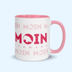 May include: White ceramic mug with a pink rim and handle. The mug features a red graphic of the island of Fehmarn with the word "Moin" in red text. The text "Fehmarn" is printed in black below the graphic. The background of the mug is covered in a repeating pattern of the word "Moin" in pink text.