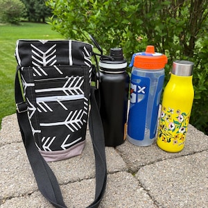 May include: A black and white arrow-patterned water bottle carrier with a black strap, next to a black water bottle, a blue and clear water bottle, and a yellow floral-patterned water bottle. The bottles are on a stone surface.