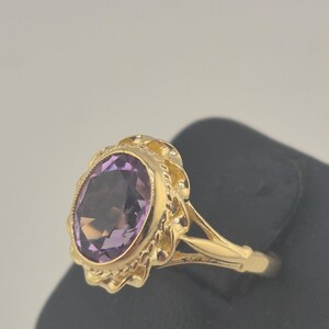 May include: A gold ring with an oval, deep purple amethyst gemstone. The gemstone is set in a decorative, scalloped bezel. The ring's band and setting are a bright, polished gold colour.