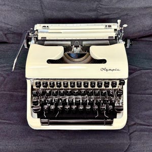 May include: A cream-colored Olympia typewriter featuring a QWERTY keyboard layout with black circular keys, silver metal accents, and a carriage assembly. The brand name Olympia is printed in a cursive script on the front right side of the casing.