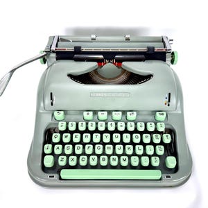 May include: A sage green Hermes Baby typewriter featuring a QWERTY keyboard with light mint green keys. The machine includes a silver carriage return lever on the left, a metal paper bail, and a central nameplate displaying the Hermes Baby brand name.