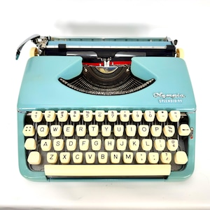 May include: A light blue Olympia Splendid 99 typewriter featuring a QWERTY keyboard with cream-colored keys. The device includes a metal carriage, a black ribbon mechanism, and the brand name Olympia Splendid 99 printed on the front casing.