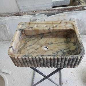 Op de afbeelding: Een witte marmeren wastafel met een gegolfde rand en drie gaten aan de bovenkant. De wastafel staat op een zwarte metalen standaard.