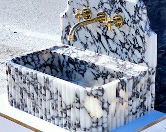 May include: A white marble sink with a gold faucet. The sink is mounted on a wall and has a decorative, fluted design. The sink is made of marble and has a white and gray color scheme.