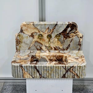 May include: A white marble sink with a brown and gold marbling pattern. The sink has a rectangular basin and a decorative, scalloped backsplash. Three gold faucets are mounted on the backsplash.