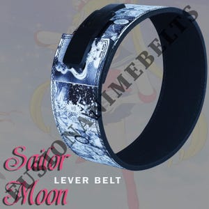 Pu&ograve; includere: Una cintura a leva di Sailor Moon in bianco e nero con fibbia a leva nera. La cintura presenta un design in stile fumetto con accenti blu e bianchi. Il testo "Sailor Moon" &egrave; visualizzato in rosa.