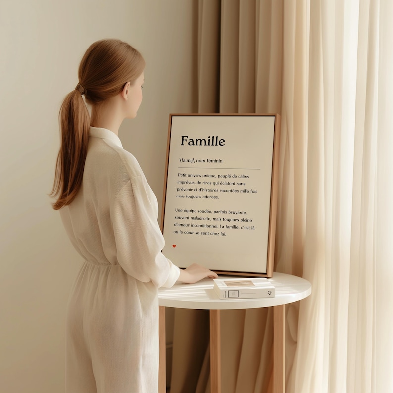 May include: A framed print with a white background and black text. The text reads "Famille" and defines the word as a unique universe filled with hugs, laughter, and stories. The print is on a white table with a wooden frame.