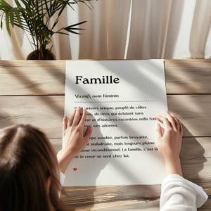 May include: A white sheet of paper with the word "Famille" written in black at the top. The word is defined in French as a unique universe, full of hugs, laughter, and stories told over and over again. The paper is on a wooden table.