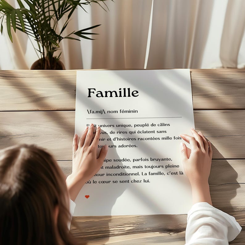 May include: A white sheet of paper with the word "Famille" written in black at the top. The word is defined in French as a unique universe, full of hugs, laughter, and stories told over and over again. The paper is on a wooden table.