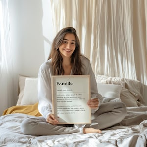 May include: A framed print with the word "Famille" in bold, black font. The print is in French and defines the word "famille" as a unique group of people who are always there for each other, even when they are imperfect. The print is held by a person sitting on a bed.