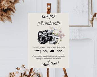 May include: A white sign with black text that says "Souriez! Photobooth" with a vintage camera illustration and a floral design. The sign also includes instructions for using the photo booth, including props like a mustache, glasses, a hat, and a mask. The text says "Seul ou à plusieurs, avec ou sans accessoires. Prenez la pose et faites sortir votre âme d'acteur. Laissez un beau souvenir aux Mariés. Have Fun!"