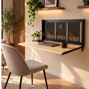May include: A wall-mounted folding desk featuring a light wood surface and a black metal back panel with a decorative cutout design of mountains, water, and a boat. A laptop and a black mug sit on the desk, which is positioned above a light-colored floor.