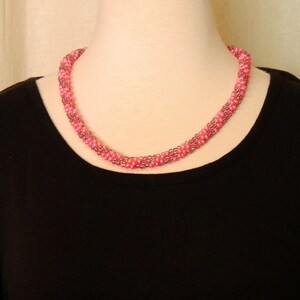 May include: A pink beaded necklace with a twisted design. The necklace is worn on a black shirt.