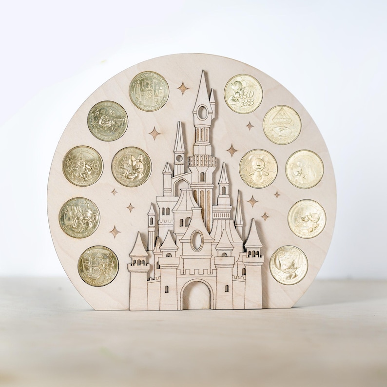 May include: A wooden cutout of a castle with a circular base. The castle is surrounded by 12 gold coins with various Disney characters on them. The background is a light wood colour.