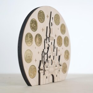 May include: A wooden cutout of a castle with a circular base. The castle is decorated with gold coins and stars. The castle is a light brown colour and the coins are a shiny gold colour.