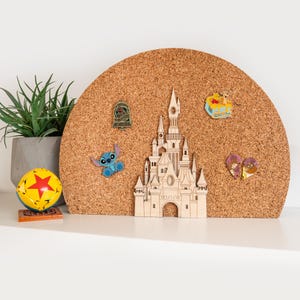 May include: A semi-circular cork display board featuring a laser-cut wooden castle centerpiece. Several character pins are attached to the board, including a blue alien, a bell-shaped pin, a yellow bear in a pot, and two heart-shaped character pins.