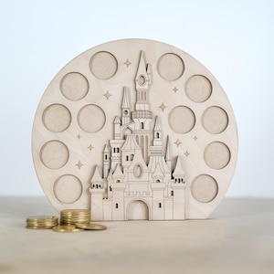 May include: A wooden coin bank shaped like a castle with a circular base and 12 coin slots. The castle is detailed with towers, windows, and a pointed roof. The bank is painted in a light wood colour.