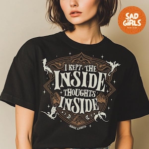May include: Black t-shirt with the text "I KEPT THE INSIDE THOUGHTS INSIDE" in white and brown. The design includes decorative elements and the text "RIDOC GAMLYN". A circular orange logo with the words "SAD GIRLS BOOK CLUB" is in the upper right corner.