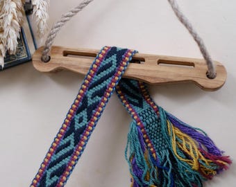 May include: A wooden wall hanging with a woven strap and tassel. The strap is blue, black, yellow, and red. The tassel is blue, green, yellow, and purple.