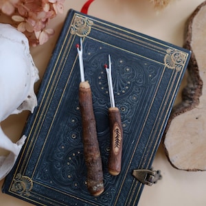 May include: Two wooden handled crochet hooks with silver metal tips. The hooks are resting on a dark blue book with gold accents.