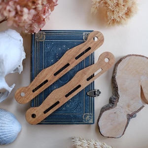 Pode incluir: Dois porta-livros de madeira com um design decorativo, colocados em cima de um livro azul e dourado com um fecho fechado. Os porta-livros estão sobre uma superfície bege claro com flores secas e um pedaço de madeira.