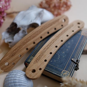 Może przedstawiać: Dwa drewniane uchwyty do pasków na książki z rzeźbionym geometrycznym wzorem i otworami do przewlekania. Paski leżą na zamkniętej książce z niebieską okładką i złotymi akcentami.