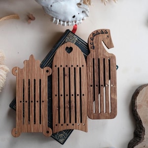 May include: Three wooden weaving tools with vertical slots and decorative shapes, including a heart and a horse head. The tools are arranged on a dark book with gold accents, with a skull and dried plants in the background.
