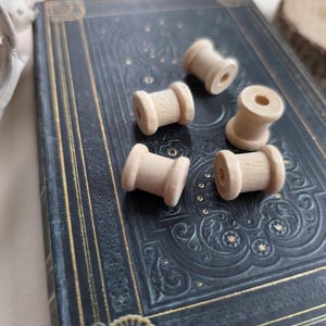 May include: Five small, unfinished wooden spools scattered on a dark blue, embossed book. The spools are cylindrical with a central hole, perfect for crafts or decorative projects. The book has gold-colored accents.