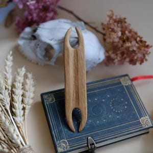 May include: A wooden knitting needle with two prongs at the top and a smooth, light brown finish. The needle is resting on a blue book with gold accents and a decorative clasp.