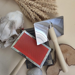 May include: Two pet grooming brushes with wooden handles and red and silver brush heads are displayed. A small skull, a seashell, a bundle of wheat, and a small book are also in the image.
