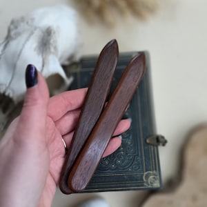 May include: Two dark brown wooden tools with rounded ends are held in a hand. The tools are smooth and appear to be made of a polished wood. The background includes a vintage book, a seashell, and a skull.