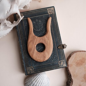 Puede incluir: Un peine de madera con un agujero central descansa sobre un libro azul oscuro en relieve. El peine es de color marrón claro y tiene un diseño curvo único. También se ven una concha blanca y un cráneo.