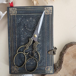 Op de afbeelding: Een donkerblauw notitieboek in antieke stijl met gouden details en een decoratieve sluiting. Een vintage schaar met gouden handvatten en zilveren bladen ligt op het notitieboek. De scène wordt aangevuld met natuurlijke elementen zoals tarwe en hout.