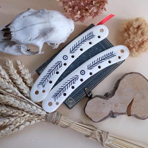 May include: Two white, curved bone hair slides with black leaf and dot designs rest on a vintage book. The image includes a skull, dried flowers, a natural sponge, and a bundle of wheat, creating a rustic, bohemian aesthetic.