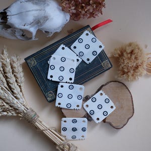 May include: Off-white square tiles with black circular patterns, arranged on a dark book, a wooden slice, and a neutral surface. The image also features dried flowers and a bundle of wheat, creating a rustic aesthetic.