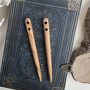 May include: Two wooden hair sticks with two holes each, resting on a dark blue, embossed book. The hair sticks are light brown with a smooth finish. The book has gold-coloured corner accents and a brass clasp.