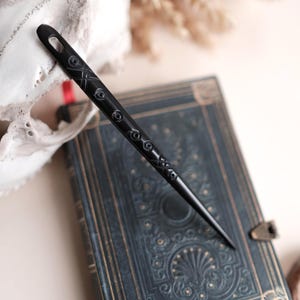 May include: A black hair pin with etched detailing, lying on a vintage-style, dark blue book with gold accents. The pin is long and slender, with a pointed end. The background includes a skull and dried plants.
