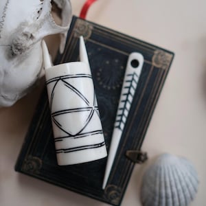 Peut inclure: Nature morte avec un objet cylindrique blanc orné de motifs géométriques noirs et un outil pointu blanc avec un motif de feuilles. Ils reposent sur un livre bleu foncé avec un ruban rouge, à côté d'un coquillage et d'un crâne.