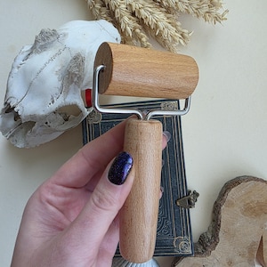 May include: A wooden rolling pin with a light brown finish and a stainless steel frame. The rolling pin is held over a vintage book, a seashell, and a piece of wood.